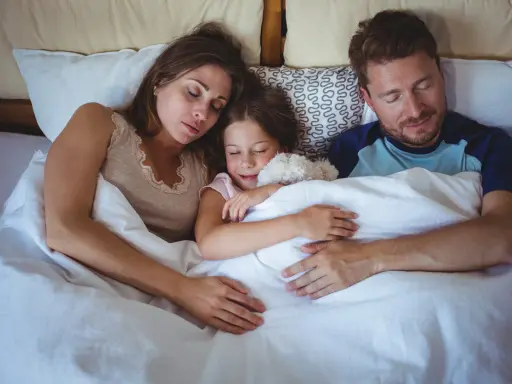 Familia durmiendo junta, la niña abraza un peluche. Un momento de paz que evoca la regresión infantil y la seguridad del hogar.