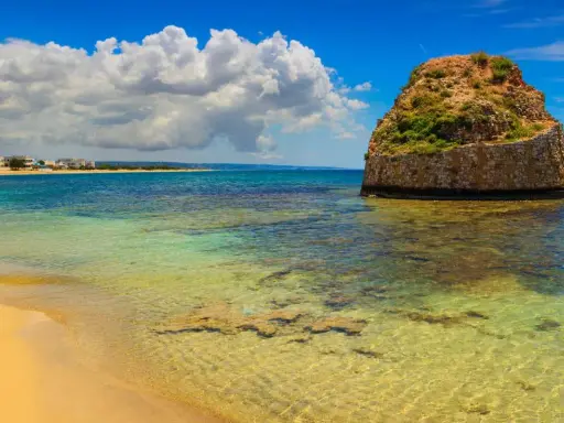 L'isola della fanciulla, una torre rotonda in pietra nel mare cristallino, con una spiaggia dorata e case in lontananza sotto un cielo azzurro con nuvole bianche.