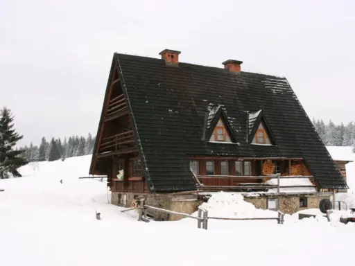 Drewniana bacówka w zimowej scenerii, otoczona śniegiem i lasem.