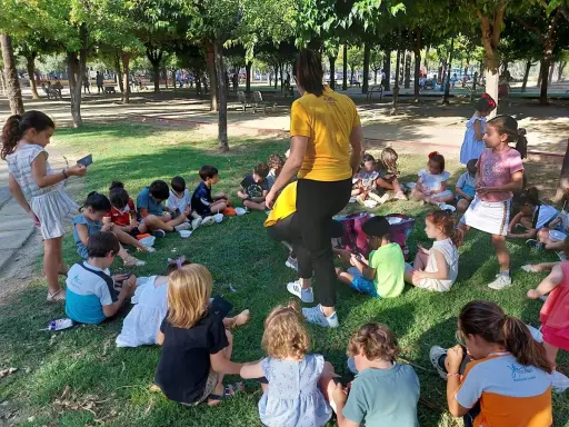 Niños sentados en el césped, participando en una gymkana para niños. Un adulto con camiseta amarilla dirige la actividad.