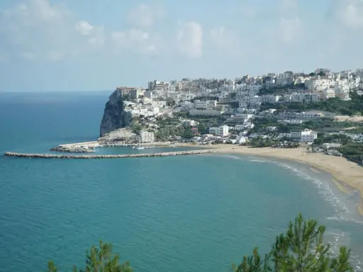 En vit stad klättrar uppför en klippa vid havet i Puglia, Italien. En strand och en hamn syns nedanför.