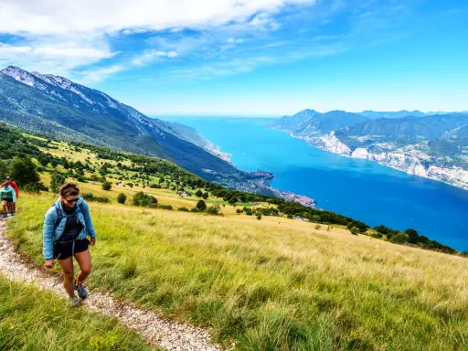 Zwei Wanderer genießen die Aussicht beim Monte Baldo Wandern. Der Weg führt über einen sonnigen Hang mit Blick auf den tiefblauen Gardasee.