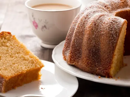 Una fetta di torta al latte soffice e dorata, cosparsa di zucchero a velo, accanto a una ciambella intera e una tazza di tè.