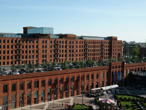Dawna fabryka w Łodzi, dziś centrum handlowe i rozrywkowe. Widać tu architekturę z czerwonej cegły, parking, ogródki kawiarniane i karuzelę, tworząc ciekawe łódzkie atrakcje.