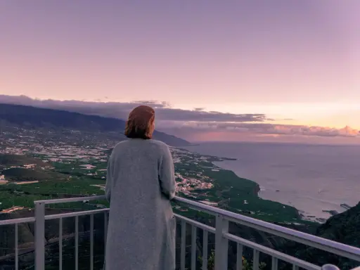 Frau blickt auf weite Landschaft, die ihr Angst vor Urlaub macht. Berge, Meer und Felder im Abendlicht.