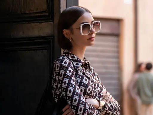 Emily in Paris, Lily Collins poses with large white sunglasses and a patterned shirt, leaning against a dark door.