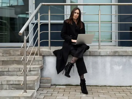 Elegancka kobieta w czarnym płaszczu i spodniach, z laptopem na kolanach, siedzi na schodach. Stylowe czarne płaszcze to podstawa wielu udanych stylizacji.