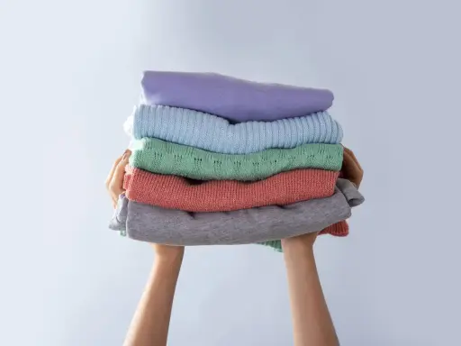 Manos sostienen una pila de ropa doblada en tonos pastel. Consejos para saber como evitar que una prenda destiña.