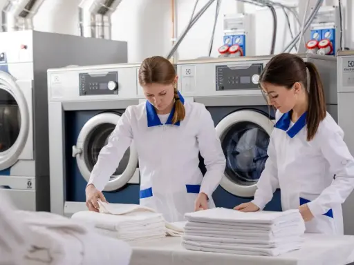 Pracownice hotelowej pralni składają czyste ręczniki. W tle widać profesjonalne pralki i suszarki.