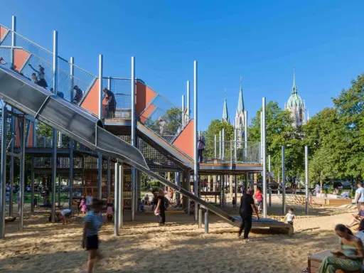 Un día perfecto de **munich con niños** en un parque infantil moderno con toboganes y estructuras de juego, con torres de iglesias al fondo.