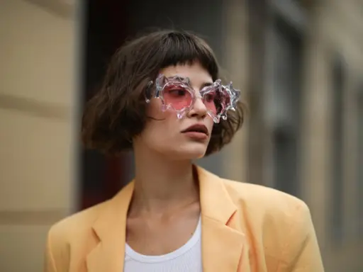 Frau mit gestuftem Bob mit Pony und auffälliger Brille mit rosa Gläsern, trägt einen hellgelben Blazer.