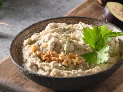 En skål med krämig baba ganoush, toppad med valnötter och persilja. Bredvid ligger skivade auberginer och ett vitlöksklyfta.
