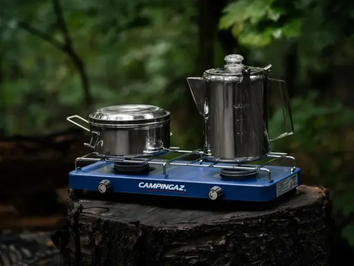 Niebieska kuchenka turystyczna Campingaz z dwoma palnikami, na której stoją metalowy garnek i kawiarka, gotowa do przygotowania posiłku w lesie.