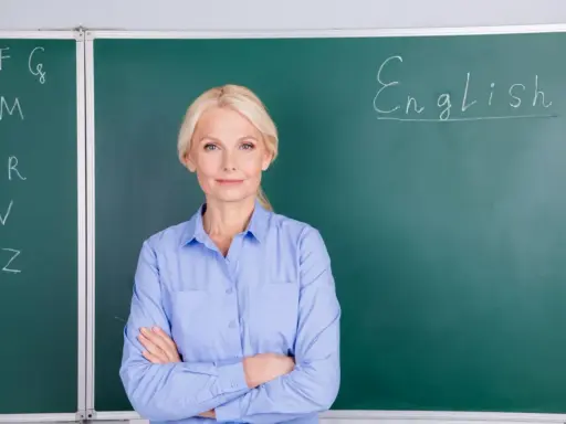 Nauczycielka angielskiego z uśmiechem, stojąca przed tablicą z napisem "English". Marzy o wcześniejszej emeryturze dla nauczycieli.