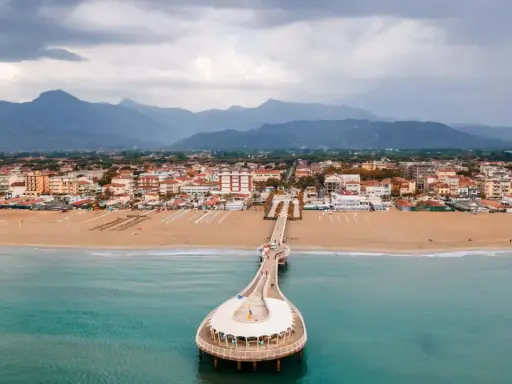 Un molo si estende su un mare azzurro, con una spiaggia dorata e una città costiera sullo sfondo. La Toscana offre un mare bello e panorami indimenticabili.