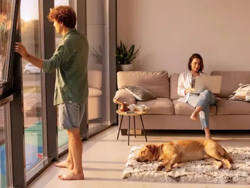 Hombre abre ventana, mujer trabaja en laptop en sofá, perro duerme. El ruido exterior, 40 dB es mucho ruido, interrumpe la paz.
