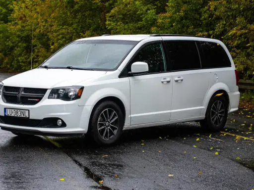 Biały Dodge Grand Caravan na mokrej drodze, otoczony jesienną zielenią.