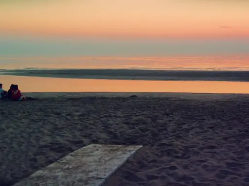 Alba a Rimini: una coppia seduta sulla sabbia guarda il mare calmo e il cielo colorato.