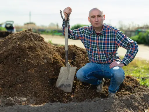 Un homme en chemise à carreaux et jean, pelle à la main, près d'un tas de tourbe blonde. Il est prêt pour son utilisation au jardin.
