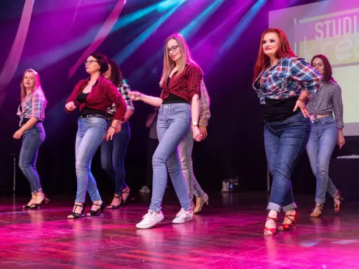 Grupa kobiet w kratę i jeansach tańczy na scenie. Występ latino solo dla początkujących.