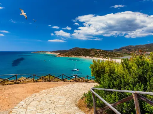 Vista panoramica di una baia con acque turchesi, spiagge affollate e montagne verdi. Un gabbiano vola nel cielo azzurro sopra Quartu e dintorni.