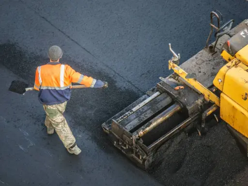 Arbeiter schaufelt heißes Bitumen auf eine Straße, während ein Asphaltfertiger das Material verteilt.