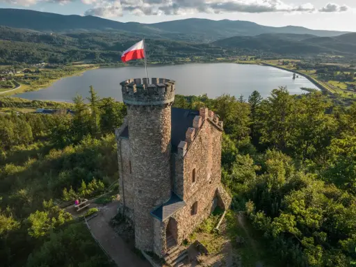 Kamienna wieża z polską flagą na tle jeziora i gór. Wspaniałe **cieplice atrakcje** dla miłośników historii i przyrody.