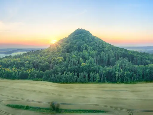 Wschód słońca nad krainą wygasłych wulkanów. Zielone wzgórze porośnięte lasem, pola uprawne i łagodnie pofałdowany krajobraz w oddali.