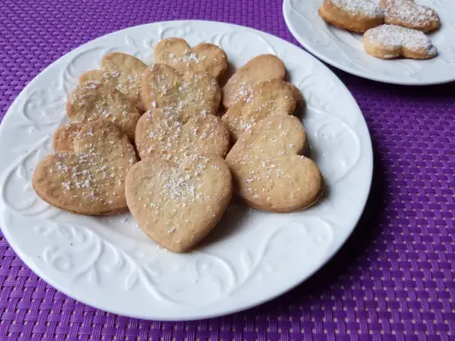 Stos ciasteczek cytrynowych w kształcie serc, posypanych cukrem pudrem, na białym talerzu.