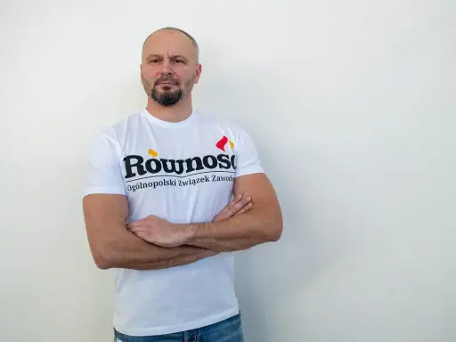 Mężczyzna w koszulce z napisem "Równość Ogólnopolski związek zawodowy" stoi z rękami skrzyżowanymi na piersi.