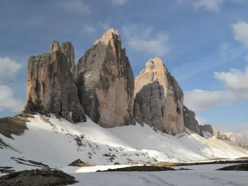 Majestatyczne **Tre Cime** w Dolomitach, ośnieżone zbocza i błękitne niebo z chmurami.