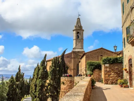 Un borgo nei comuni Val d'Orcia: un campanile svetta tra cipressi e case antiche, con un bar all'aperto.
