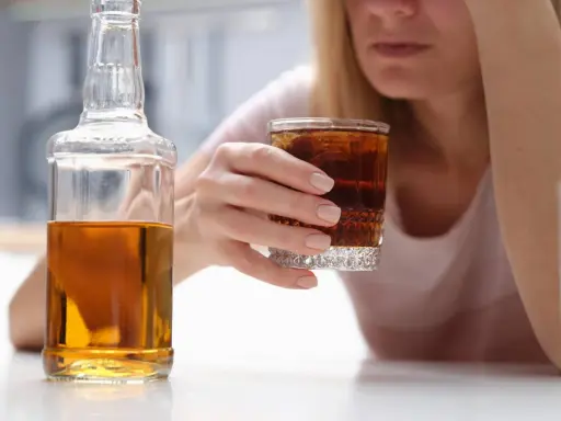 Kobieta z kieliszkiem drinka i butelką alkoholu. W tle widać jej twarz, która wyraża smutek. To może być wszywka alkoholowa.