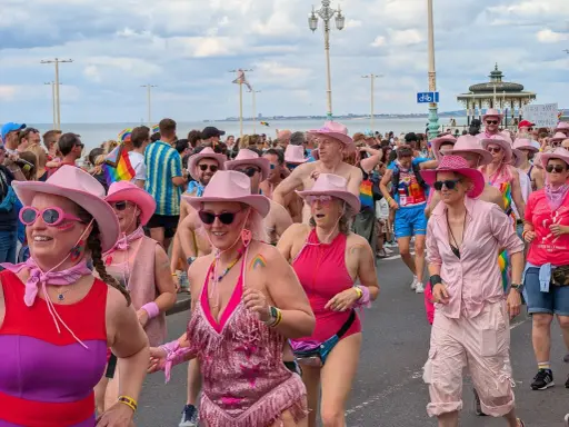 A vibrant Brighton gay pride parade, with many participants wearing pink cowboy hats and rainbow-themed attire, celebrating joyfully by the sea.