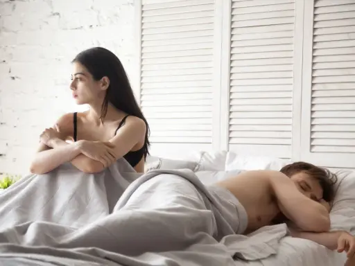 Kobieta patrzy w bok, zmartwiona, podczas gdy mężczyzna śpi. W związku brakuje pociągu seksualnego.