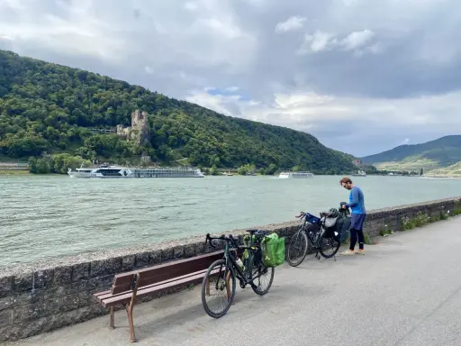 Mann packt Gepäck für eine Fahrradtour am Rhein. Im Hintergrund Burg und Fluss.