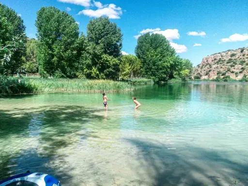 Dos niños juegan en las cristalinas aguas de las Lagunas de Ruidera, rodeados de vegetación y un paisaje montañoso.