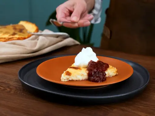 En bit krämig ostkaka med keso, toppad med vispgrädde och sylt. Serveras på en orange tallrik.