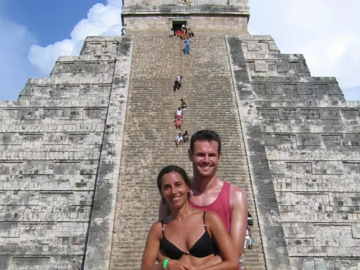 Coppia sorridente davanti alla piramide di Kukulkan, con turisti che salgono le scale del chichen itza interno.