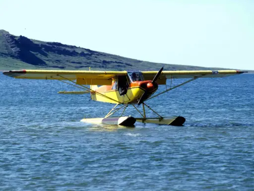 Gelbe Piper Super Cub mit Schwimmern auf ruhigem Wasser, im Hintergrund eine grüne Hügelkette.