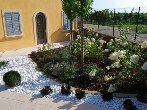 Ingresso esterno di una villetta con ghiaia bianca, piante fiorite e un vialetto. Belle idee per l'esterno della casa.