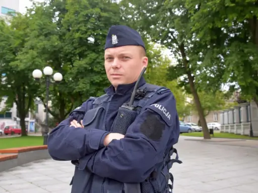 Policjant w granatowym mundurze, w sprawie umundurowania policjantów, stoi na tle zieleni.