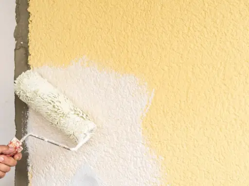 Hand streicht mit einer Farbrolle über eine raue, gelbe Wand. Der Putz wird gerade weiß gestrichen.