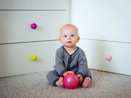 7 miesięczne dziecko z niebieskimi oczami siedzi na dywanie, trzymając w rączkach różową piłeczkę.