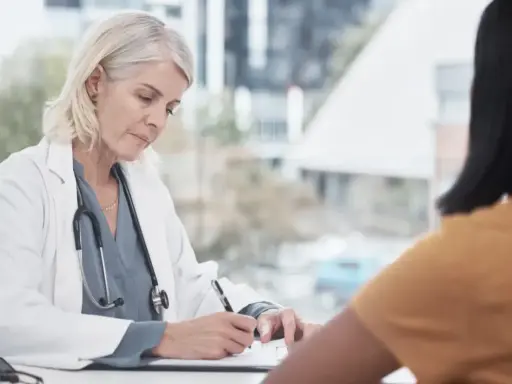 Lekarka w białym kitlu z receptą w ręku, pacjentka w żółtej bluzce. Wizyta u lekarza, może l4 na jeden dzień.