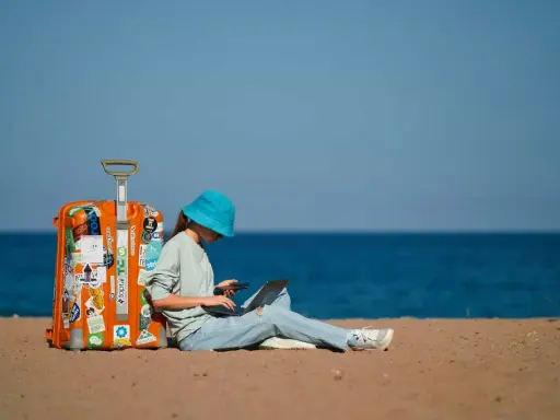 Dziewczyna w niebieskim kapeluszu siedzi na plaży z walizką, laptopem i telefonem, planując, jak tanio podróżować.
