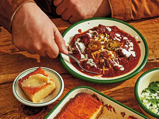 En hand tar en sked ur en skål med mustig texas chili, toppad med jalapeños och gräddfil. Bredvid ligger gyllene majsbröd.
