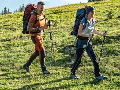 Trzy osoby wędrują po zielonym zboczu. Mężczyzna i kobieta w wygodnych, sportowych spodniach w góry, z plecakami i kijkami trekkingowymi.