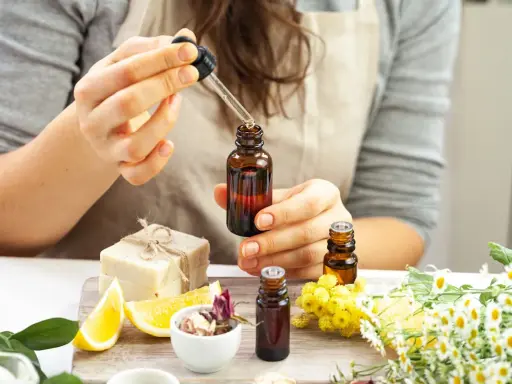 Une personne prépare un parfum huile essentielle, ajoutant une goutte d'ambre dans un flacon. Des fleurs, des citrons et du savon sont disposés autour.