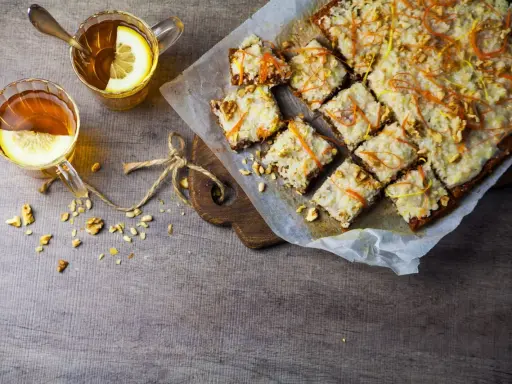 En skiva vegansk morotskaka med glasyr och nötter, serverad med två koppar te med citron.